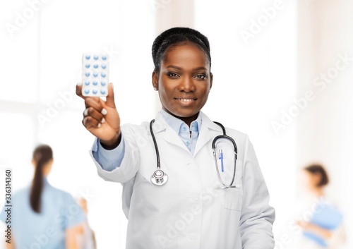 medicine, profession and healthcare concept - happy smiling african american female doctor or in white coat with pills and stethoscope over hospital staff on background photo