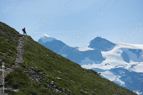 Tourist with backpack standing on path and enjoying beautiful mountains scenery on background. Mountain hiking, men reaching peak in sunny summer day. Sport tourism in Alps.
