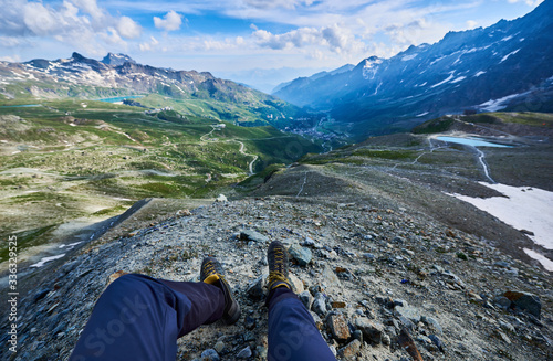 Male legs in hiking boots resting on hillside in mountain valley. Beautiful view of mountains and hills under cloudy sky with man hiker feet on foreground. Concept of travelling, hiking and alpinism.
