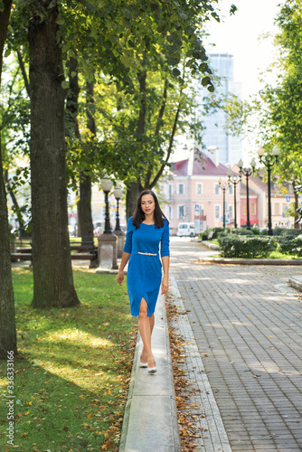 young handsome brunette business woman in blue dress walking on the parapet in the city park