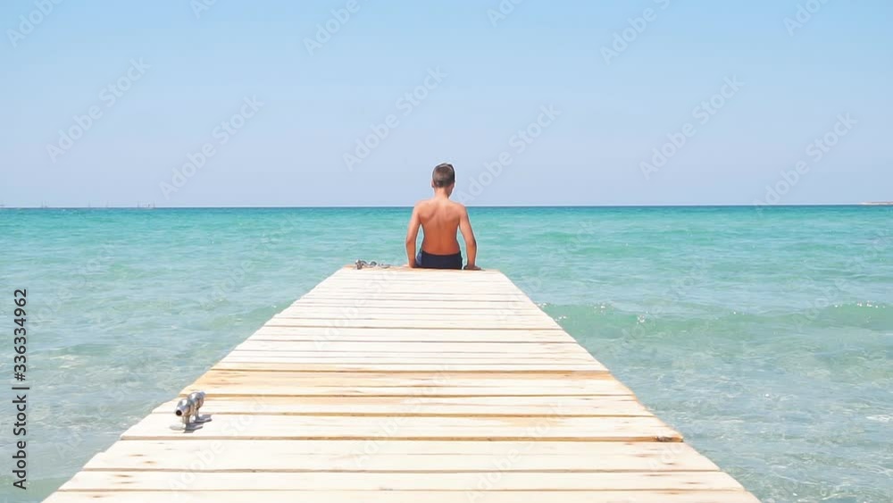 Teenager sits on the edge of wooden pier against the backdrop of beautiful azure sea, slow-motion video. Teenager sits on the edge of wooden pier against the backdrop of beautiful azure sea, slow-motion video.