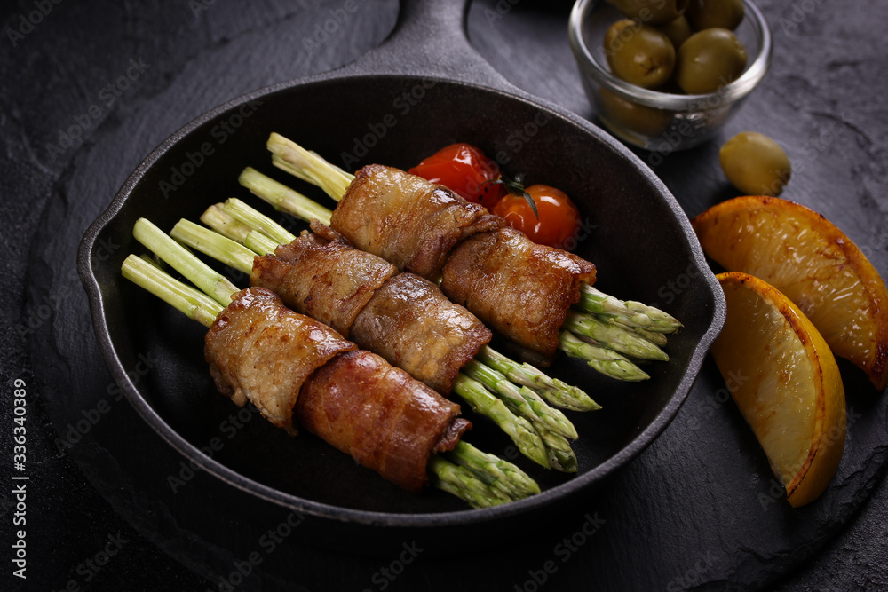 Fried bacon and asparagus in a black cast iron pan on a black background. Fried tomatoes and lemon, green olives and ketchup. Background image, copy space Fried bacon and asparagus in a black cast iron pan on a black background. Fried tomatoes and lemon, green olives and ketchup. Background image, copy space