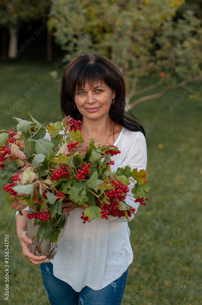 vertical close-up portrait of a fifty-year-old woman with a bouquet of viburnum in her hands who stands in the middle of the garden on green grass at sunset