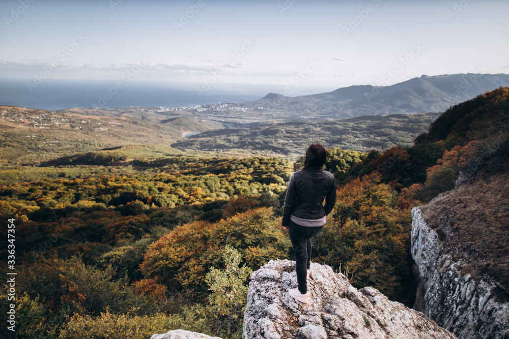 caucasian woman in dark jeans and a dark jacket stands on the edge of a cliff, and looks at the landscape of an autumn valley going to the sea against a blue sky