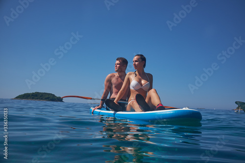 Young and beautiful couple of lovers sitting on a surfboard in the open ocean on a background of sky. © ZoomTeam