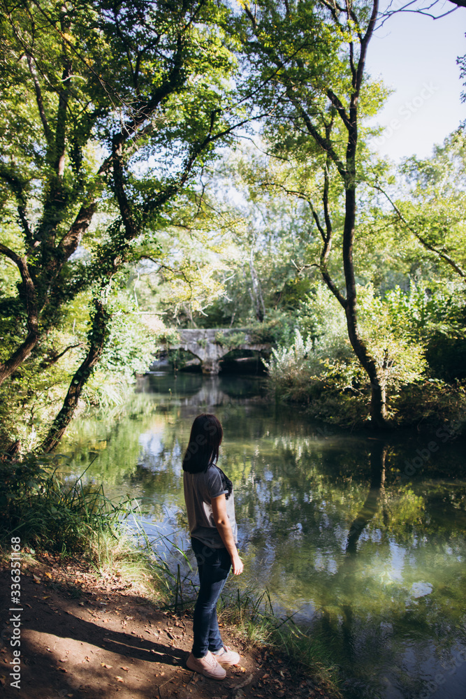girl with dark hair in a gray t-shirt stands on the background of old dark stone bridge across the river. bridge with reflection in the river among green trees.