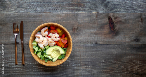 Healthy salad with shrimp, avocado, cucumbers, and tomatoes. Balanced lunch in bowl. Wooden background with copy space. Top view.