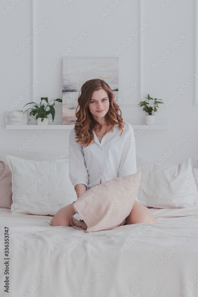 young beautiful brunette in white shirt sitting on the bed holding a pillow in her hands and smiling