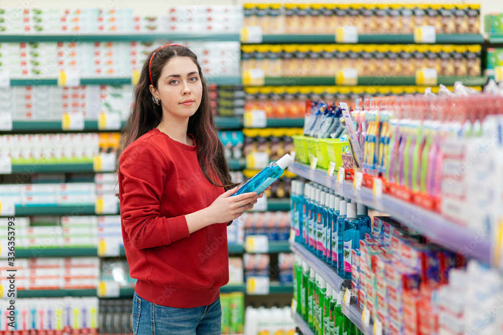 A young Caucasian woman in a red sweater holds a bottle of shampoo and smiles tenderly. In the background, shelves with cosmetics blurred. The concept of buying cosmetics A young Caucasian woman in a red sweater holds a bottle of shampoo and smiles tenderly. In the background, shelves with cosmetics blurred. The concept of buying cosmetics