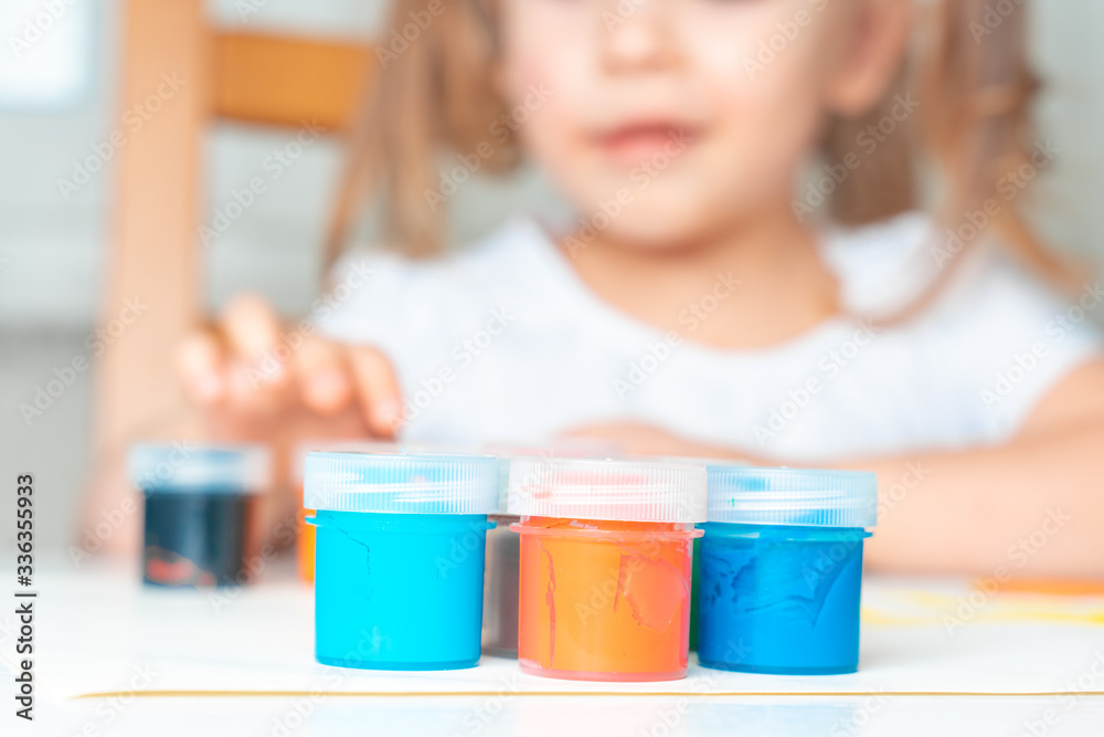 set of gouache in jars placed on a table for children's creativity. In the background a child in blur. activities with the child in isolation and quarantine in connection with the coronavirus set of gouache in jars placed on a table for children's creativity. In the background a child in blur. activities with the child in isolation and quarantine in connection with the coronavirus