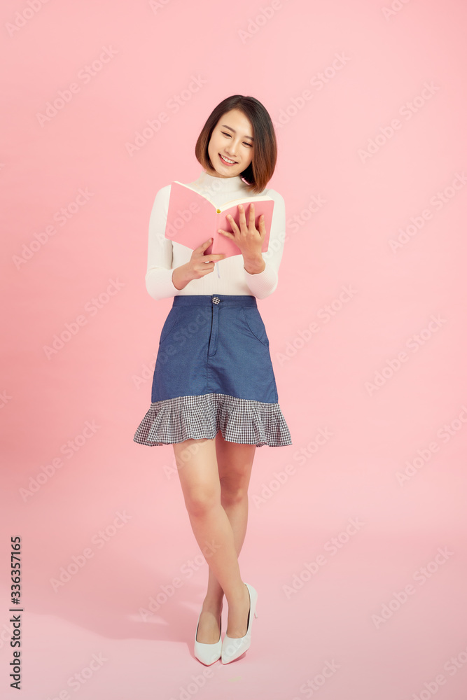 Full length portrait of a smiling beautiful Asian woman student holding book isolated on a pink background Full length portrait of a smiling beautiful Asian woman student holding book isolated on a pink background