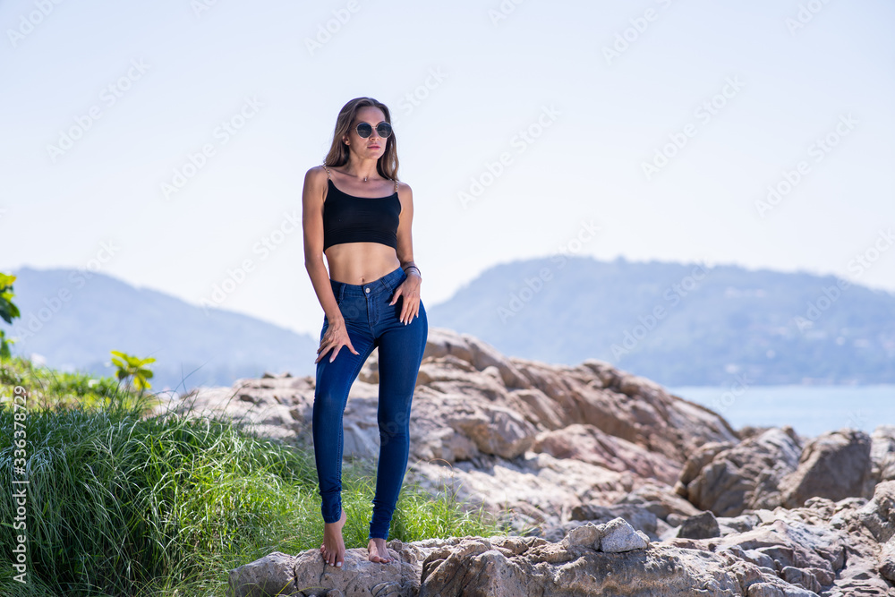 Naklejka premium Fashion shooting with female model stand on beach stone in tropical place, behind sea, dark blue jeans, black top, sunglasses, summer time.