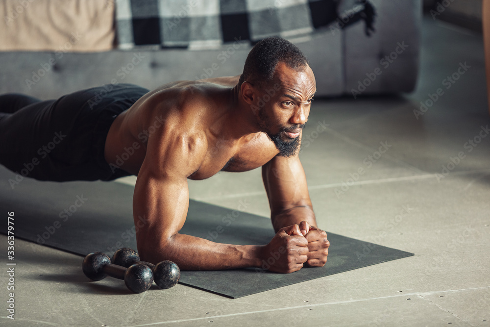 Obraz premium African-american man teaching at home online courses of fitness, aerobic, sporty lifestyle while being quarantine. Getting active while isolated, wellness, movement concept. Training, plank.