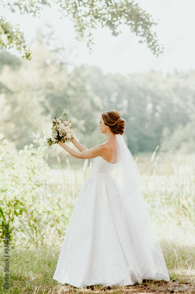 The bride in a white wedding dress and white veil is holding a bouquet of peonies in the background of a green park