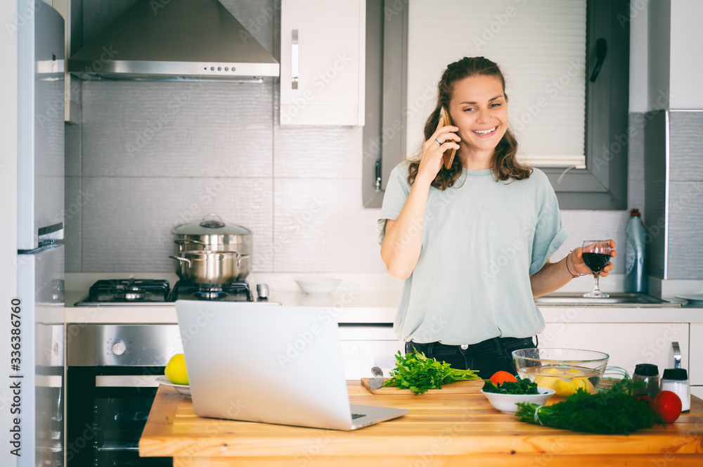 Happy beautiful young woman is cooking in the kitchen at home and talking on the smartphone with a glass of wine in her hand, communication education, cooking, lifestyle concept Happy beautiful young woman is cooking in the kitchen at home and talking on the smartphone with a glass of wine in her hand, communication education, cooking, lifestyle concept