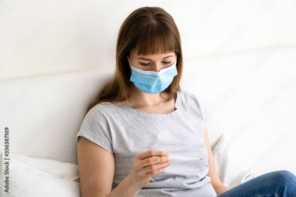 Girl in protective mask with thermometer is sitting on bed