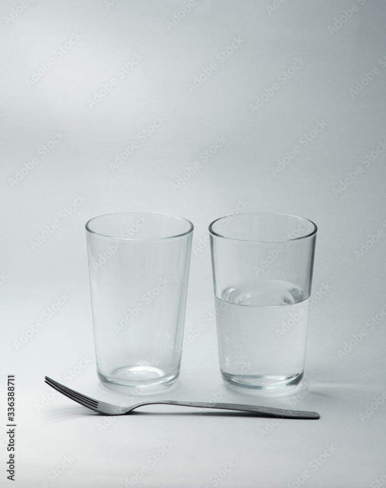 A glass cup half full of water and empty glass stands on a light background. Close-up. Copy space A glass cup half full of water and empty glass stands on a light background. Close-up. Copy space