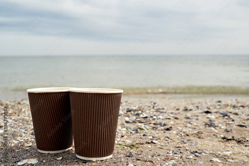 Two disposable paper cups of coffee standing on sand against blue sea, copy space. Take-out coffee Two disposable paper cups of coffee standing on sand against blue sea, copy space. Take-out coffee