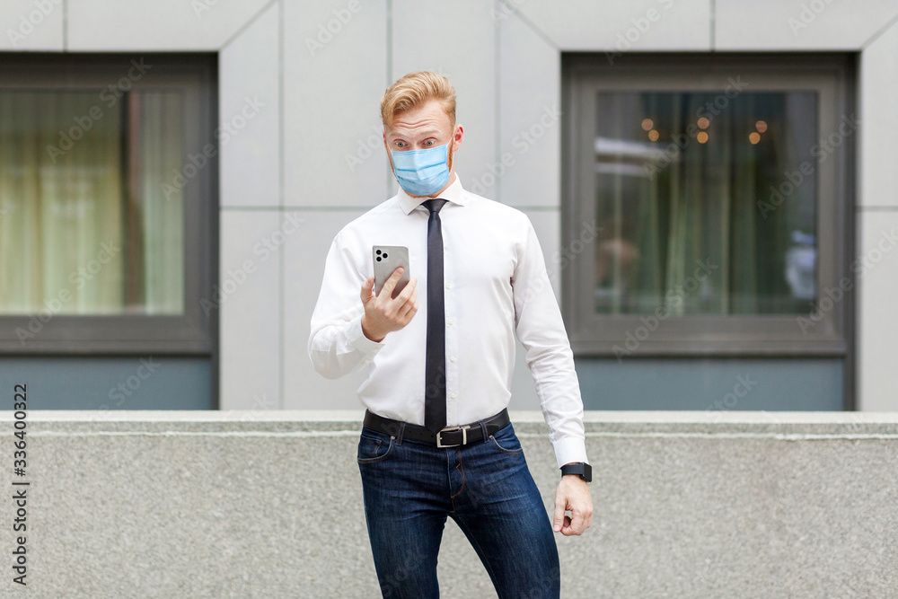 Young shocked man with surgical medical mask looking at smart phone, reading news with shocked face. Outdoor shot. business, working and health care concept.