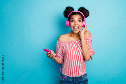 Photo of shocked funky dark skin lady hold telephone read good news listen music modern earphones wear trendy red white striped shirt off-shoulders jeans isolated blue color background