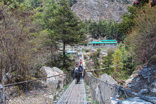 One of many pedestrian suspension bridges with waving prayer flags in Himalayas on the way to Everest Base Camp in Nepal. Hikers walks to the other side of the bridge towards buildings.