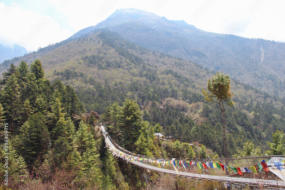 One of many pedestrian suspension bridges with waving prayer flags in Himalayas on the way to Everest Base Camp in Nepal. Walking Sherpa porter is visible on the other side of footbridge.