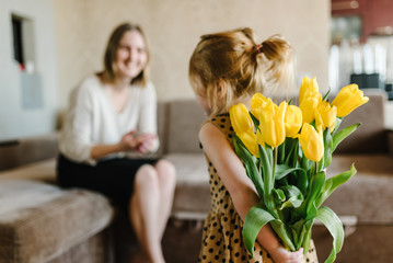 Cute little girl greeting mother and gives her a bouquet of flowers tulips at home. Mother's day concept. Mom and daughter smiling. Happy family holiday and togetherness. Back view.