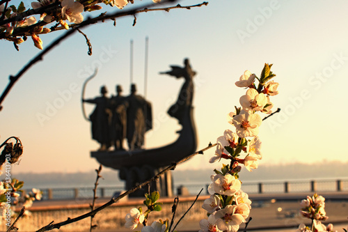 White blossom flowers on tree branch in sunny spring morning. Blurred silhouette of Monument to legendary founders of Kyiv: Kiy, Schek, Khoryv and their sister Lybid in the background. Kyiv, Ukraine photo