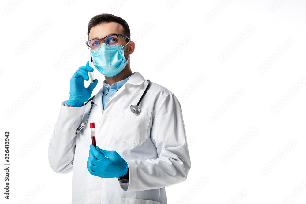 doctor in medical mask and white coat talking on smartphone while holding test tube with blood sample isolated on white