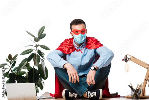 man in medical mask and superhero costume sitting on table isolated on white