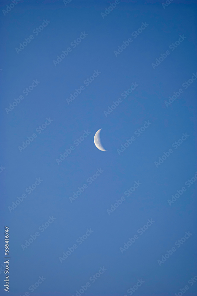 A slim sliver of the silvery moon set on an expanse of clear and cloudless blue sky.