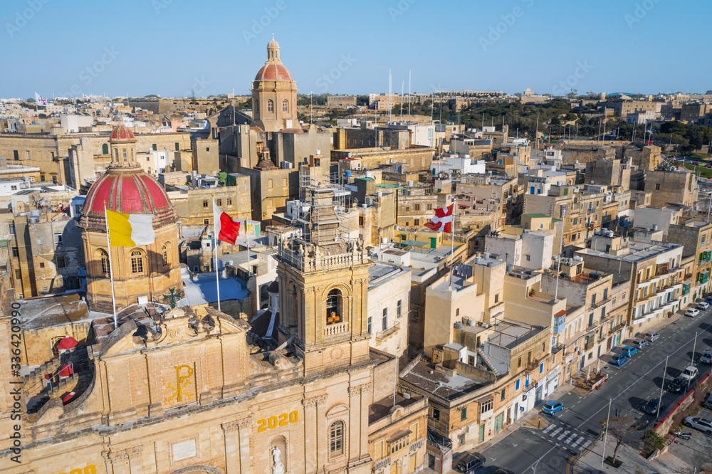 Aerial top view of Birgu city. Main catholic church, red dome and many flags on the roofs. Clear blue sky. Malta island