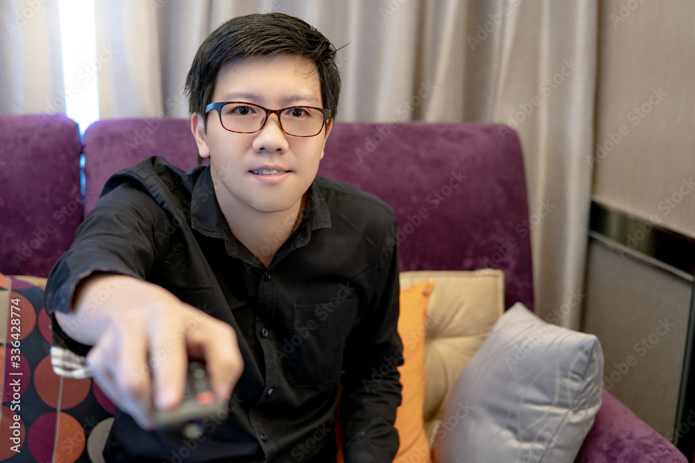 Asian happy man using remote controller while watching TV sitting on sofa in living room. Domestic life during quarantine. Stay home with entertainment media and live streaming. Asian happy man using remote controller while watching TV sitting on sofa in living room. Domestic life during quarantine. Stay home with entertainment media and live streaming.