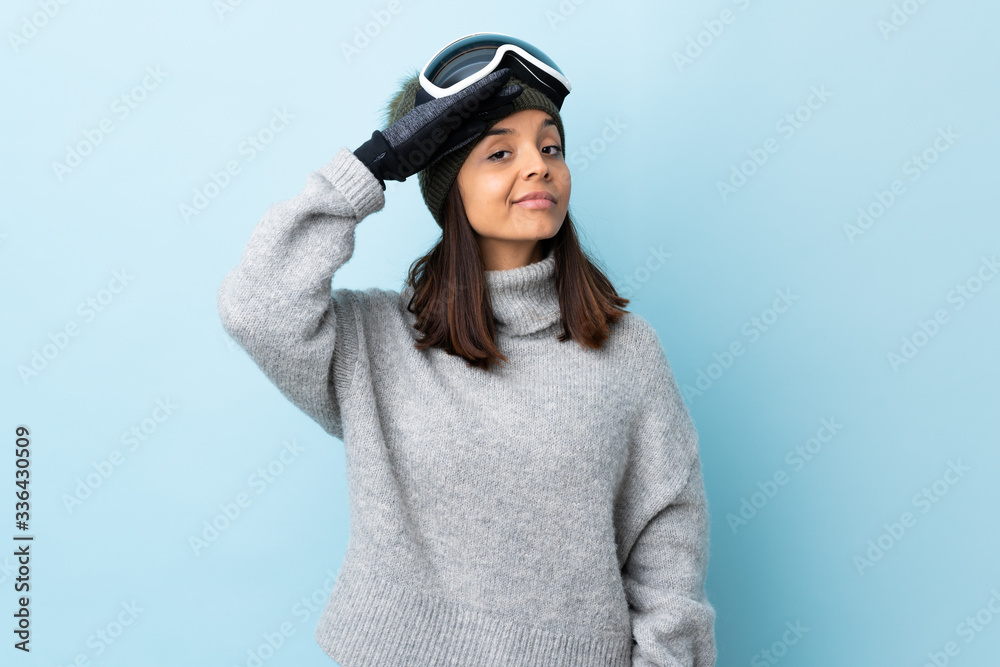 Mixed race skier girl with snowboarding glasses over isolated blue background saluting with hand with happy expression. Mixed race skier girl with snowboarding glasses over isolated blue background saluting with hand with happy expression.