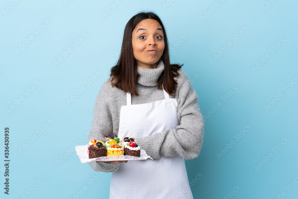 Pastry chef holding a big cake over isolated blue background making doubts gesture while lifting the shoulders. Pastry chef holding a big cake over isolated blue background making doubts gesture while lifting the shoulders.