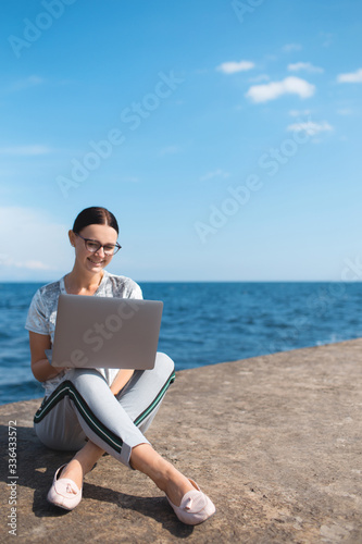 Happy young woman sitting with a laptop by the sea. Webinar online teaching concept. Online work.