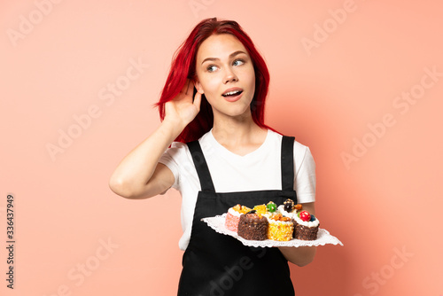 Pastry chef holding a muffins isolated on pink background listening to something by putting hand on the ear
