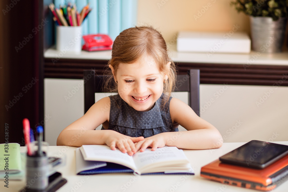 Little blonde girl doing homework at home at the table. The child is home-schooled. A girl with light hair performs a task online using a laptop and tablet computer. Read books. Little blonde girl doing homework at home at the table. The child is home-schooled. A girl with light hair performs a task online using a laptop and tablet computer. Read books.