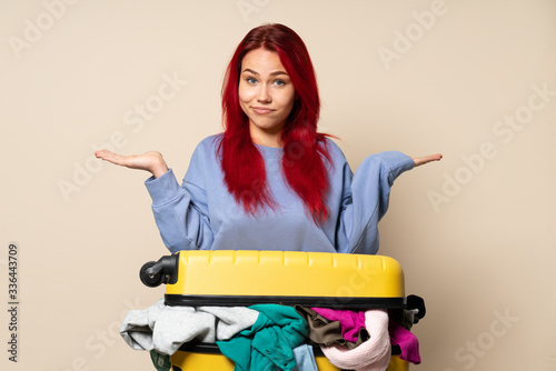 Traveler girl with a suitcase full of clothes isolated on beige background having doubts while raising hands photo