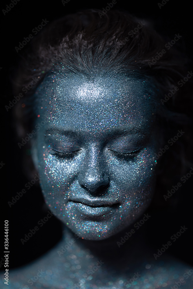 girl with a blue face in sparkles close-up against a black wall 