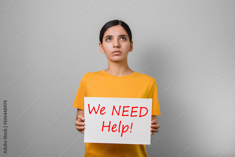 we need help. dramatic portrait of a young woman in yellow t-shirt holding a placard and looking up asking for help against grey background we need help. dramatic portrait of a young woman in yellow t-shirt holding a placard and looking up asking for help against grey background