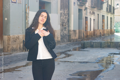 Portrait of caucasian bussiness woman model with closed eyes, keeps hands on chest, in the city, white shirt and black jacket, dark air. Place for your text in copy space. © Sergio Barceló