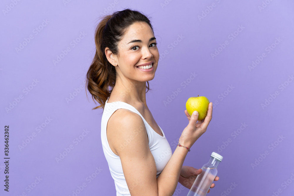 Woman over isolated purple background with an apple and with a bottle of water Woman over isolated purple background with an apple and with a bottle of water