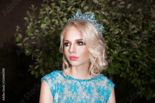 Beauty blond curly hair young woman portrait outside green park background blue crystal crown and blue dress looking away to the side