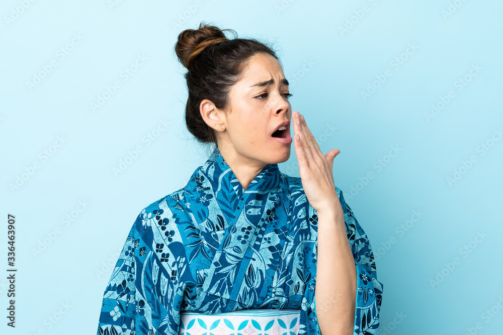 Woman wearing kimono over isolated background yawning and covering wide open mouth with hand Woman wearing kimono over isolated background yawning and covering wide open mouth with hand