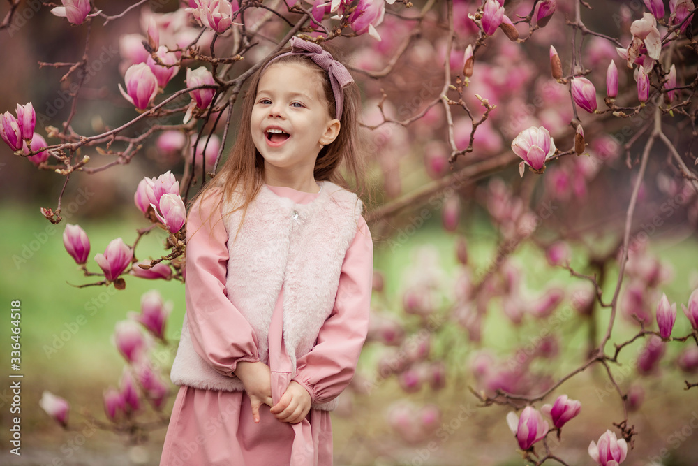 Portrait of a cute cheerful girl in a pink dress wearing a fur vest who laughs on a walk in the spring in a park under a flowering pink magnolia tree