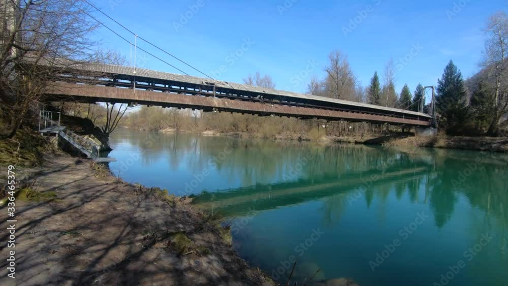Left side moving with right pan view of a wooden foot suspension bridge crossing a calm green river on a sunny winter day Left side moving with right pan view of a wooden foot suspension bridge crossing a calm green river on a sunny winter day