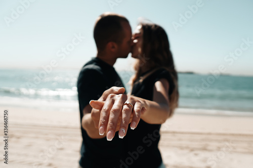 Fresh engaged couple enjoying a romantic engagement day on the beach during a sunny day. Close up on her ring.