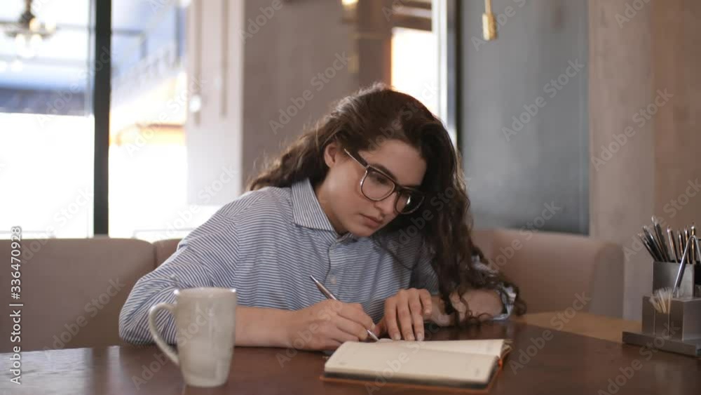 Young beautiful businesswoman in eyeglasses sitting at cafe table with coffee cup and writing down in notepad Young beautiful businesswoman in eyeglasses sitting at cafe table with coffee cup and writing down in notepad