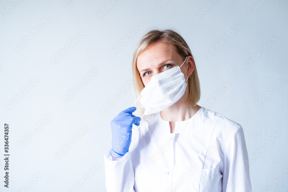 Blonde female doctor surgeon in white uniform, protective blue gloves putting on medical mask over grey. Healthcare, stay home. Copyspace. Blonde female doctor surgeon in white uniform, protective blue gloves putting on medical mask over grey. Healthcare, stay home. Copyspace.